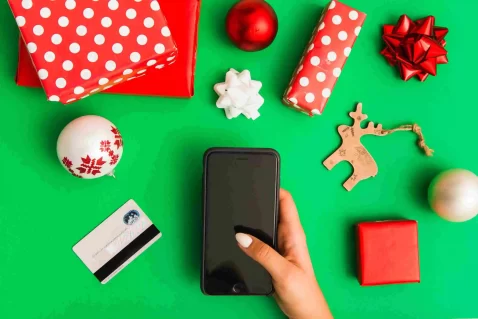 Mão segurando celular sobre mesa verde com presentes de Natal, enfeites e cartão de crédito ao redor, representando compras online de fim de ano