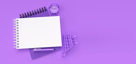 Caderno em branco sobre fundo lilás com caneta, relógio e mini carrinho de compras, simbolizando estratégias de e-commerce para 2026