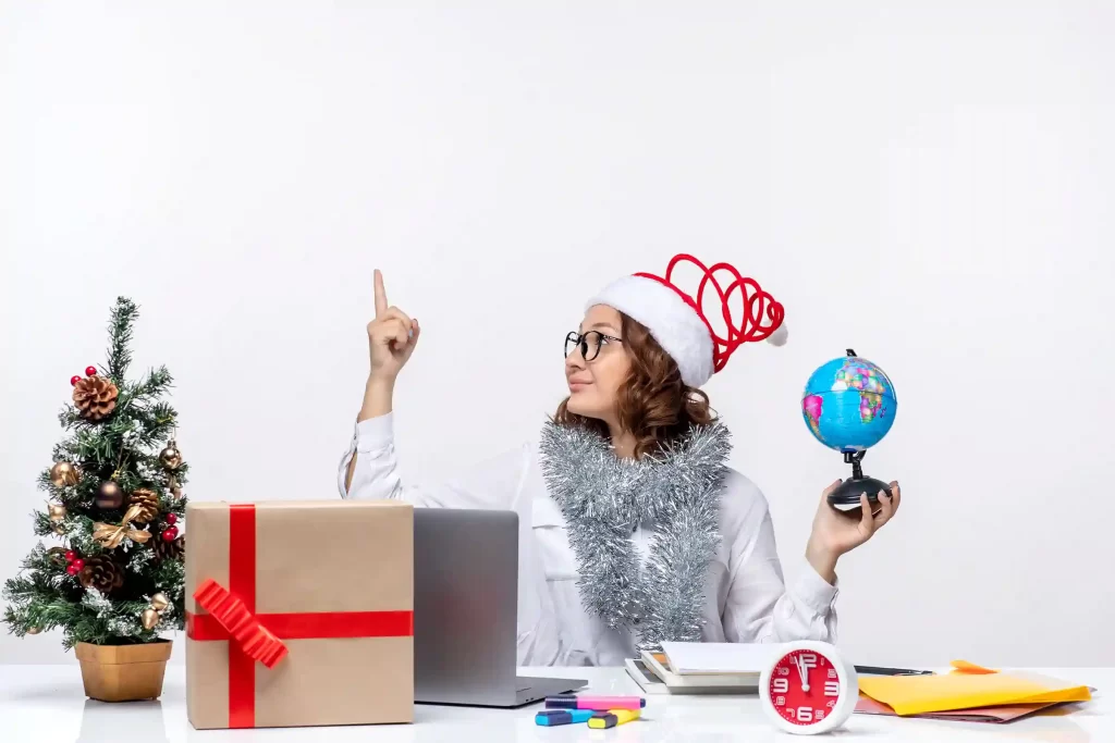 Mulher com gorro de Natal e enfeites festivos trabalhando em uma mesa com notebook, globo terrestre e presente decorado, simbolizando a preparação da loja para o fim do ano
