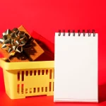 Cesta amarela com presentes embrulhados e laço dourado ao lado de um bloco de anotações em branco, sobre fundo vermelho, representando o planejamento de vendas de fim de ano