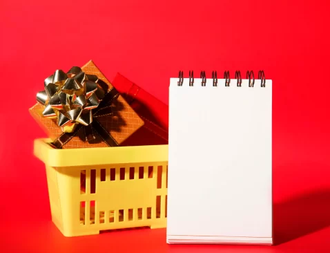 Cesta amarela com presentes embrulhados e laço dourado ao lado de um bloco de anotações em branco, sobre fundo vermelho, representando o planejamento de vendas de fim de ano