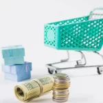 Carrinho de compras em fundo branco com pilha de moedas e notas enroladas em destaque, simbolizando análise de lucro e aplicação da regra 80/20 no MeLi