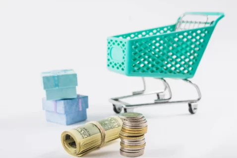 Carrinho de compras em fundo branco com pilha de moedas e notas enroladas em destaque, simbolizando análise de lucro e aplicação da regra 80/20 no MeLi