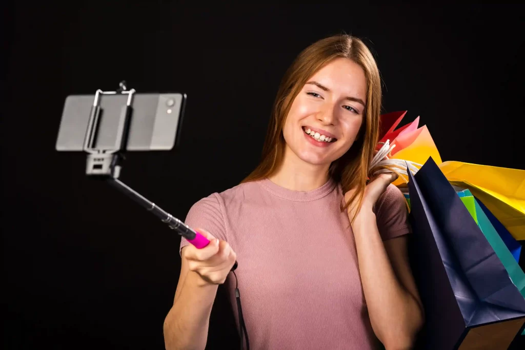 Mulher sorridente gravando vídeo com bastão de selfie e várias sacolas coloridas de compras, representando criadora de conteúdo promovendo produtos no YouTube Shopping