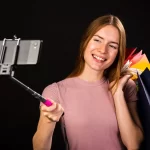 Mulher sorridente gravando vídeo com bastão de selfie e várias sacolas coloridas de compras, representando criadora de conteúdo promovendo produtos no YouTube Shopping