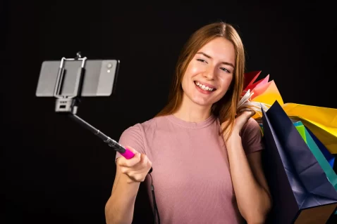Mulher sorridente gravando vídeo com bastão de selfie e várias sacolas coloridas de compras, representando criadora de conteúdo promovendo produtos no YouTube Shopping