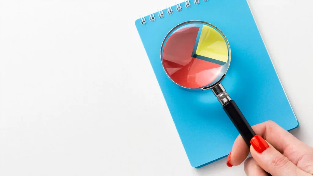 Mão segurando uma lupa sobre um caderno azul com gráfico em forma de pizza colorido, representando o estudo de dados em uma análise de concorrência