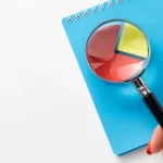 Mão segurando uma lupa sobre um caderno azul com gráfico em forma de pizza colorido, representando o estudo de dados em uma análise de concorrência