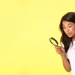 Mulher observando algo com uma lupa em frente a um fundo amarelo, representando a análise detalhada na busca de produtos para vender.