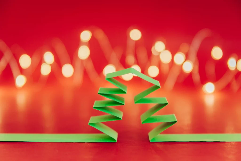 Árvore de Natal estilizada feita de tiras dobradas de papel verde, com luzes desfocadas ao fundo sobre um cenário vermelho