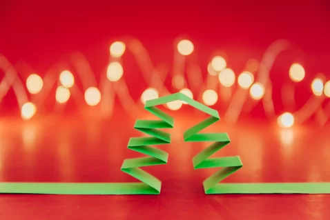 Árvore de Natal estilizada feita de tiras dobradas de papel verde, com luzes desfocadas ao fundo sobre um cenário vermelho