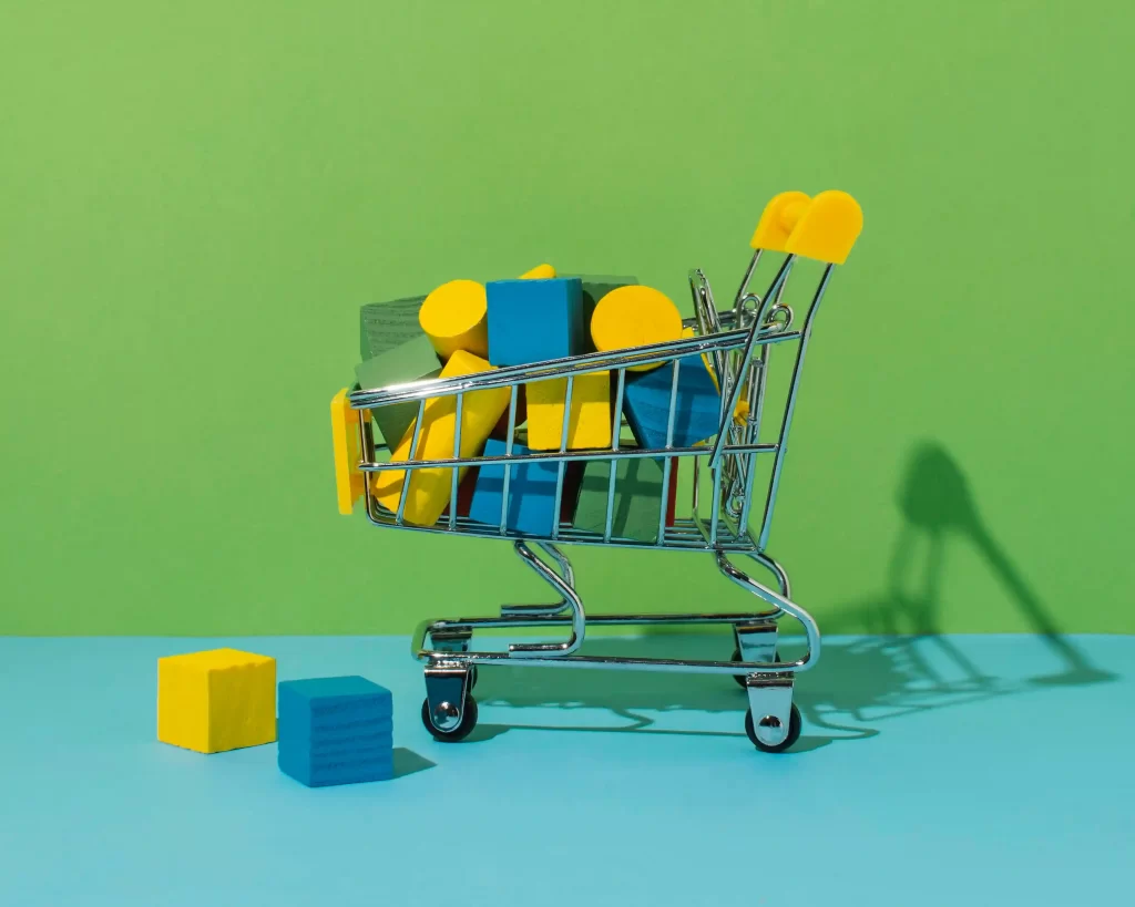 Carrinho de compras em miniatura cheio de blocos coloridos sobre fundo verde e azul, simbolizando a diversidade de subcategorias e a redução de riscos de monopolização no mercado