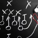 Quadro negro com desenhos de tática em giz e apito pendurado, representando o planejamento para prever reações da concorrência