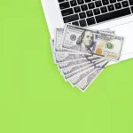 Notas de cem dólares espalhadas parcialmente sobre o teclado de um laptop branco, posicionados sobre uma superfície verde