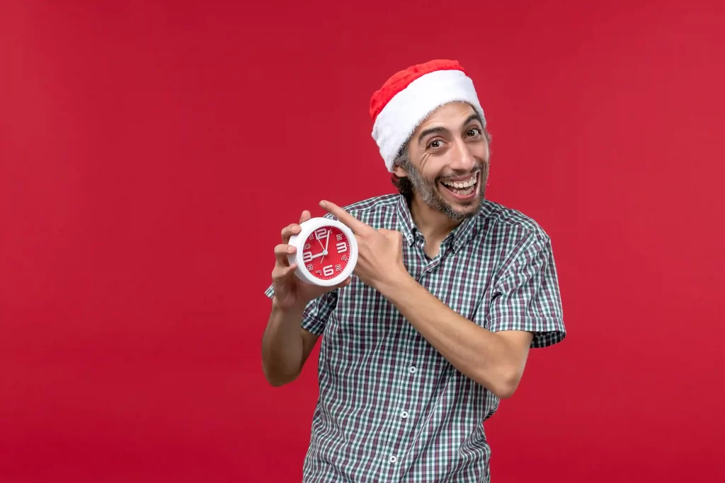 Homem sorridente usando gorro de Papai Noel e apontando para um relógio vermelho, simbolizando o clima de pressa nas vendas de última hora do Natal 2025