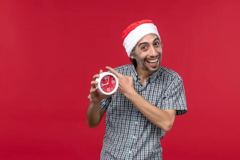 Homem sorridente usando gorro de Papai Noel e apontando para um relógio vermelho, simbolizando o clima de pressa nas vendas de última hora do Natal 2025