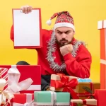 Homem com gorro de Papai Noel e enfeite de pisca-pisca no pescoço segura uma prancheta em branco enquanto está cercado por várias caixas de presentes coloridos sobre um fundo amarelo