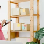 Mulher usando tablet para anotar dados enquanto observa prateleiras com caixas etiquetadas, simbolizando o acompanhamento de produtos de venda recorrente em um pequeno negócio