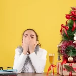 Mulher sentada à mesa com as mãos cobrindo o rosto, aparentando cansaço ou frustração, ao lado de uma pequena árvore de Natal decorada com laços vermelhos