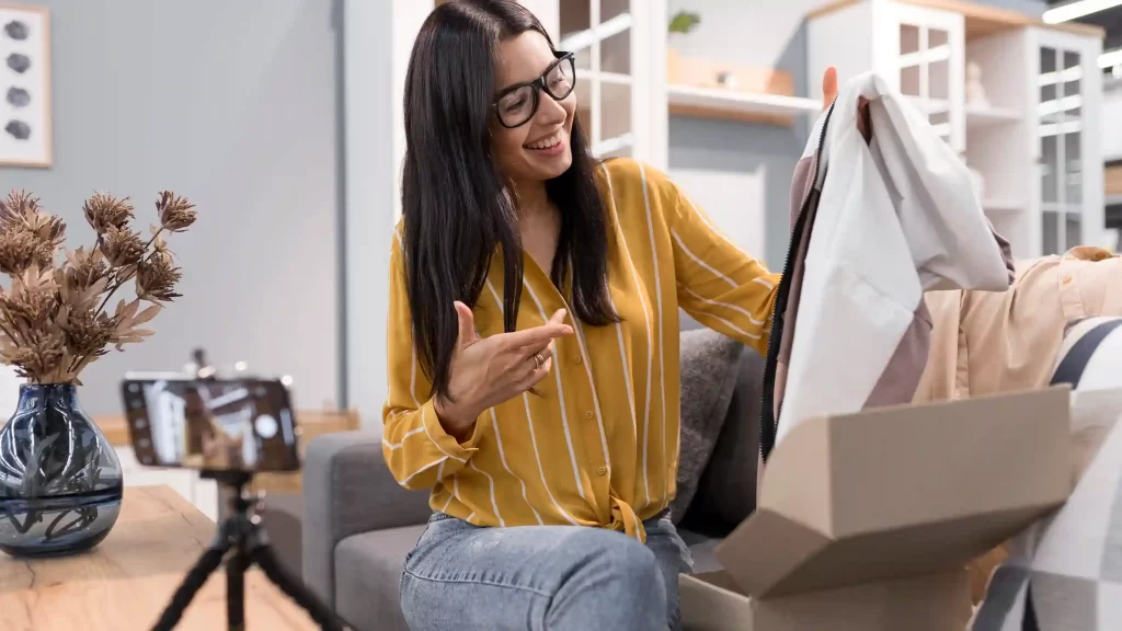Mulher sentada em uma sala, sorrindo e mostrando uma peça de roupa para a câmera do celular posicionada em um tripé