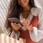 Mulher segurando sacolas de compras enquanto usa o celular, sorrindo e interagindo com a tela