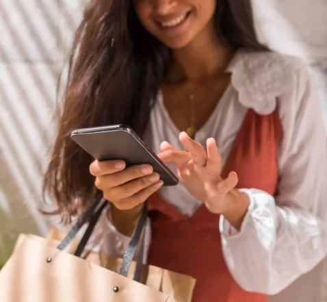 Mulher segurando sacolas de compras enquanto usa o celular, sorrindo e interagindo com a tela