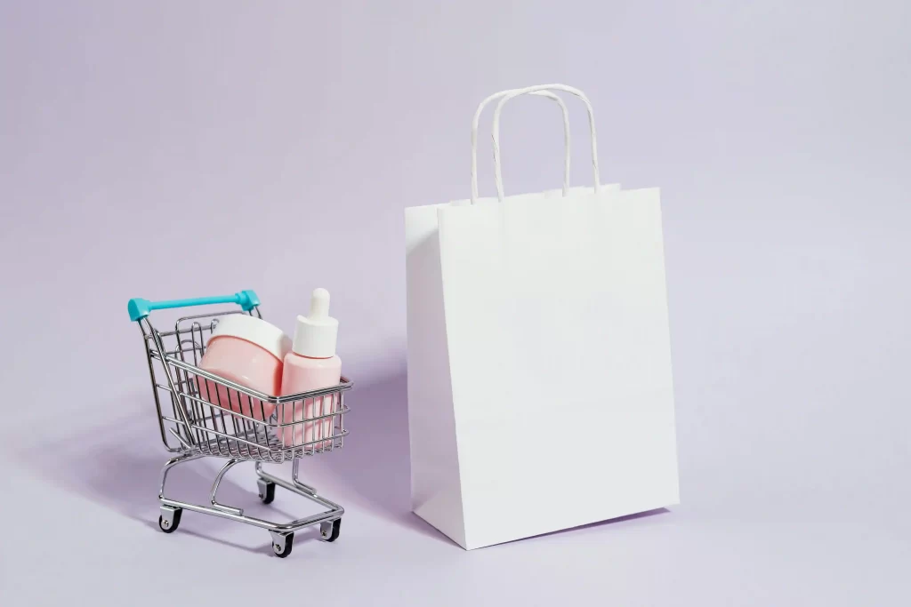 Carrinho de compras em miniatura com produtos de skincare rosa ao lado de uma sacola branca sobre fundo lilás