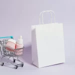 Carrinho de compras em miniatura com produtos de skincare rosa ao lado de uma sacola branca sobre fundo lilás