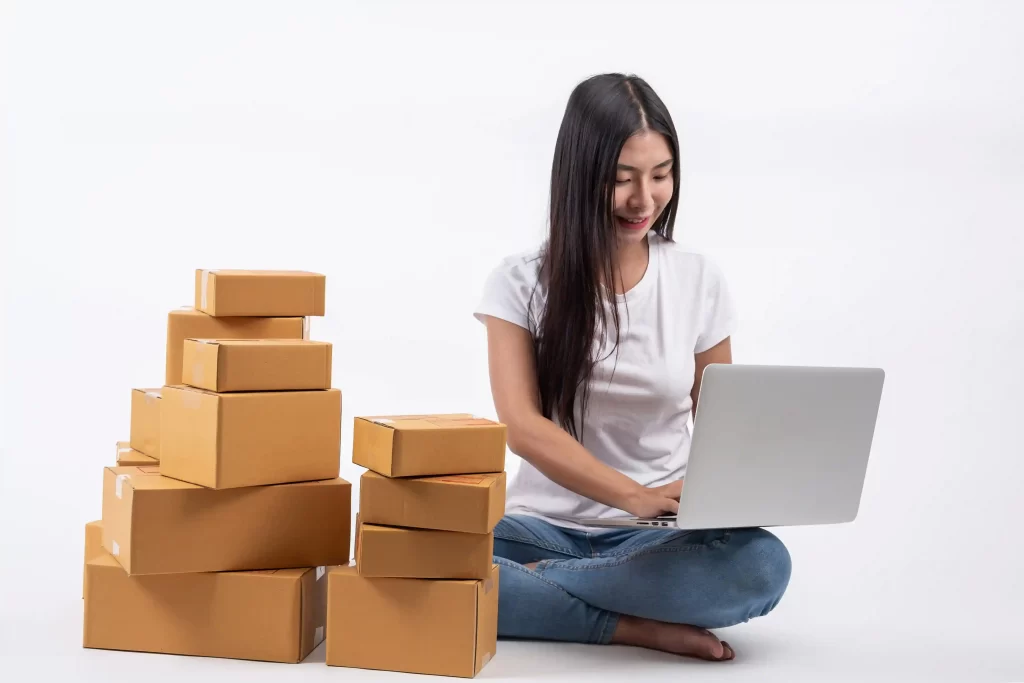 Mulher sentada no chão, com as pernas cruzadas, usando camiseta branca e jeans, trabalhando em um notebook prateado ao lado de várias caixas de papelão