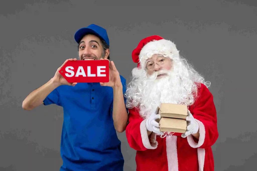 Homem sorridente vestido de uniforme azul segurando uma placa vermelha com a palavra SALE ao lado de um Papai Noel que carrega pequenas caixas de encomenda
