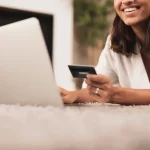 Mulher sorrindo enquanto usa notebook e segura cartão de crédito para finalizar compra online