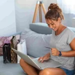 Mulher fazendo compras online no laptop com cartões e sacolas ao lado.