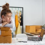 Mulher vendedora apoiada em caixas, olhando para o notebook com expressão de preocupação, enquanto produtos e pedidos se acumulam ao redor