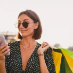 Mulher jovem usando óculos escuros segura um celular com a mão direita e carrega sacolas de compras coloridas no braço esquerdo