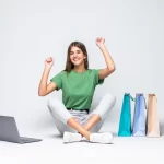Mulher jovem sentada no chão com pernas cruzadas, sorrindo e levantando os braços em comemoração; ao lado há um notebook aberto no chão e três sacolas de compras coloridas sobre fundo branco