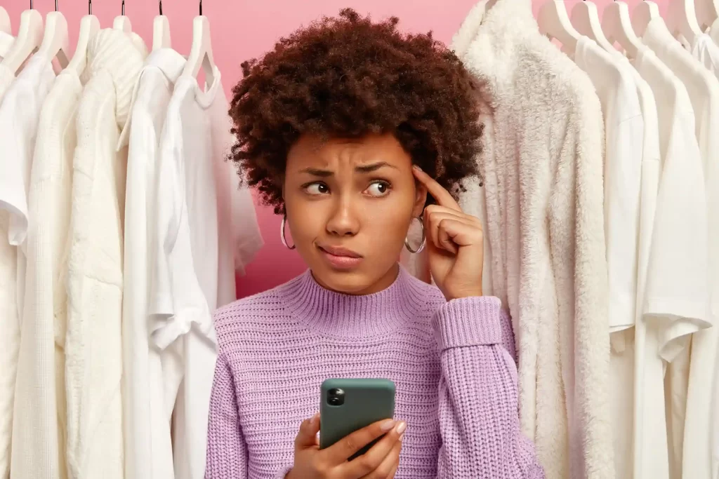Mulher de cabelo crespo curto, vestindo blusa lilás, segurando um celular e tocando a lateral da cabeça com expressão de dúvida