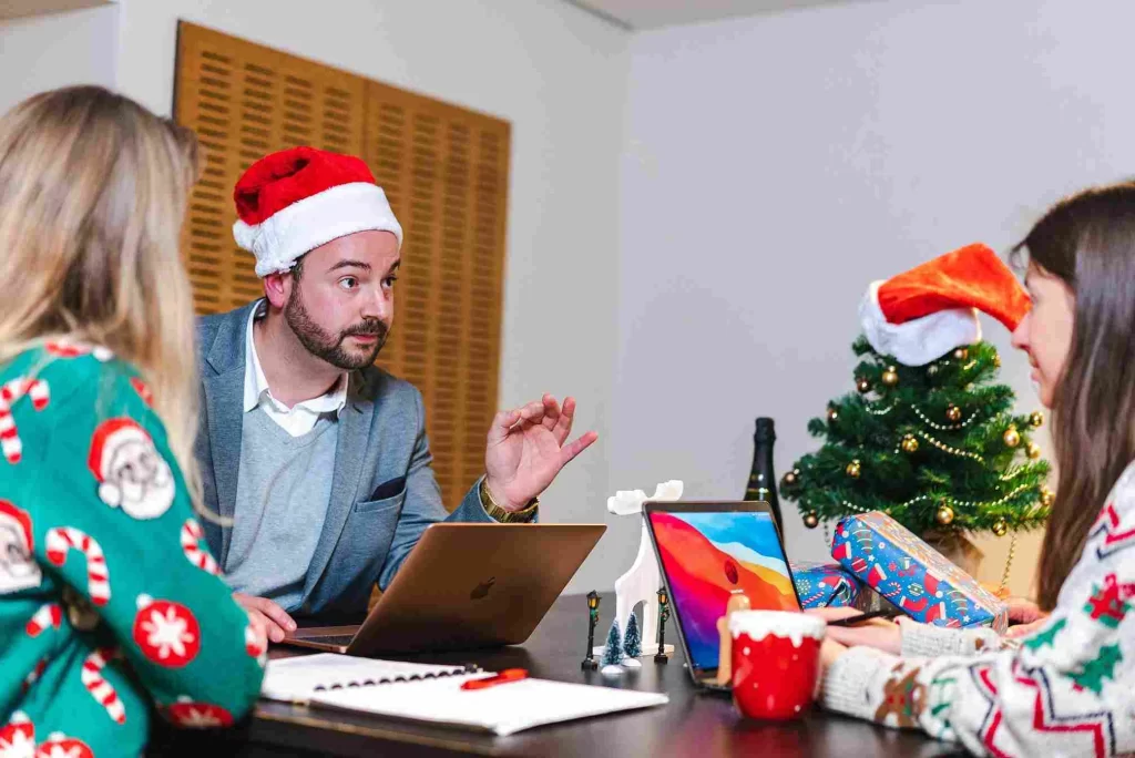 Pessoas em reunião usando gorros de Natal, analisando estratégias no notebook com árvore decorada ao fundo