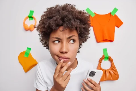 Pessoa segurando celular e olhando pensativa para roupas e acessórios colados na parede ao fundo.