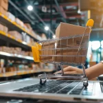 Pessoa usando um notebook em um armazém, com um carrinho de compras em miniatura contendo uma caixa de papelão sobre o teclado