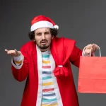 Homem vestindo roupa de Papai Noel vermelha e gorro natalino, com expressão de dúvida, segurando uma sacola de compras vermelha em uma mão e a outra mão aberta, sobre fundo cinza, representando incerteza após o Natal