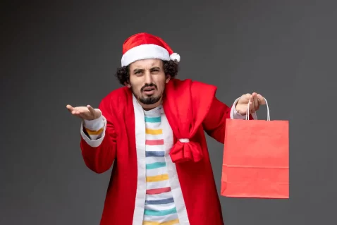 Homem vestindo roupa de Papai Noel vermelha e gorro natalino, com expressão de dúvida, segurando uma sacola de compras vermelha em uma mão e a outra mão aberta, sobre fundo cinza, representando incerteza após o Natal