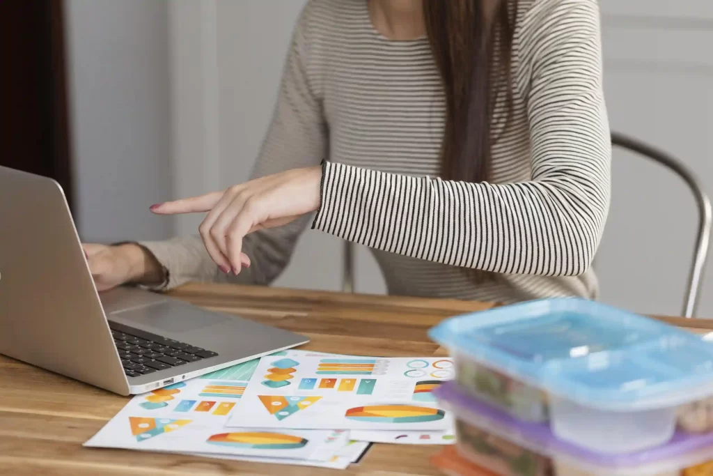 Mulher sentada à mesa usando um notebook, apontando para a tela com a mão direita, com folhas impressas de gráficos coloridos espalhadas sobre a mesa