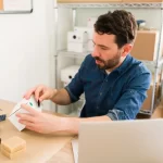 Homem em um pequeno estoque ou oficina, analisando uma caixa branca com as mãos, com um notebook aberto à sua frente