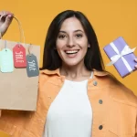 Mulher sorrindo em fundo amarelo, segurando uma sacola de compras com etiquetas escritas “SALE” e um presente roxo com laço branco nas mãos