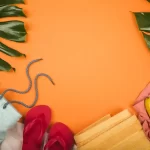 Fundo laranja visto de cima, com folhas verdes tropicais nos cantos, um par de chinelos vermelhos, toalhas dobradas em tons claros, uma sacola de tecido com cordão e um cacho de bananas à direita