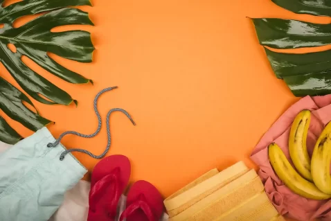 Fundo laranja visto de cima, com folhas verdes tropicais nos cantos, um par de chinelos vermelhos, toalhas dobradas em tons claros, uma sacola de tecido com cordão e um cacho de bananas à direita