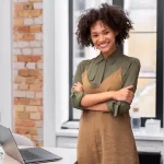 Mulher sorridente em escritório moderno, em pé ao lado de uma mesa com notebook aberto, representando confiança e planejamento para empreender no e-commerce