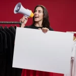 Mulher em pé dentro de uma loja de roupas, segurando um megafone e um cartaz branco em branco, com araras de roupas pretas e brancas ao fundo.