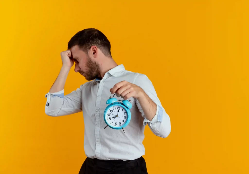 Homem vestindo camisa clara em frente a um fundo amarelo, segurando um despertador azul com a mão direita enquanto apoia a cabeça com a mão esquerda
