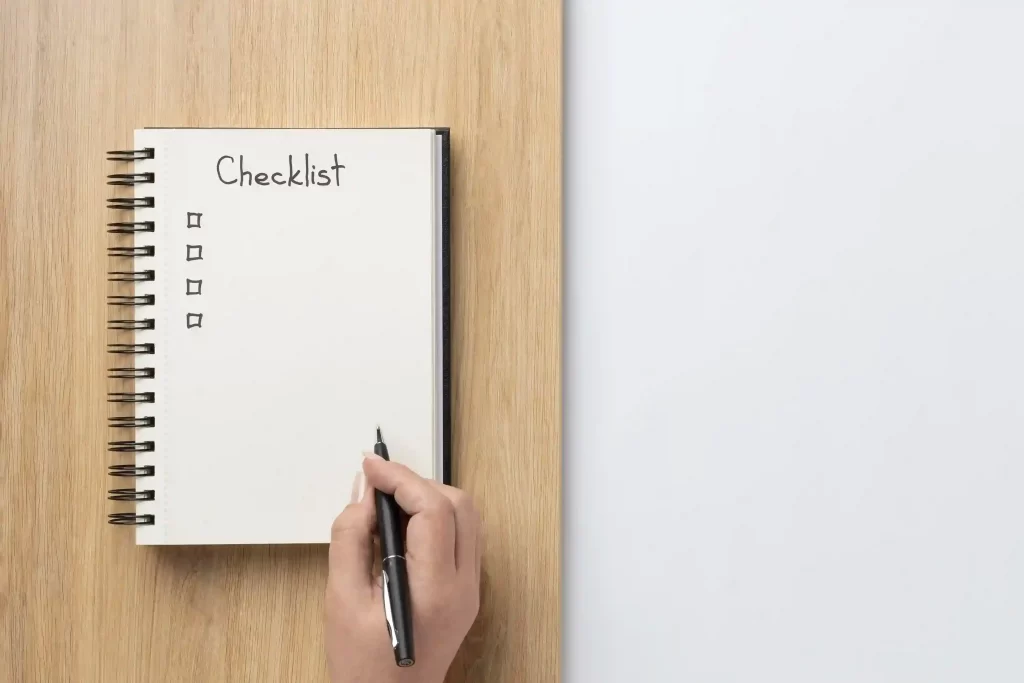 Mão segurando uma caneta sobre um caderno espiral aberto, com a palavra “Checklist” escrita no topo da página e quatro caixas de marcação vazias, apoiado sobre uma mesa de madeira clara.